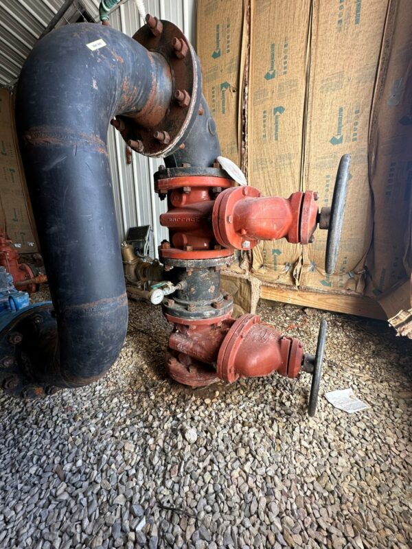 Large black and red industrial pipes with two valve wheels, installed on a gravel floor inside a metal building—ideal for supporting operations on a cattle ranch or hunting property, with insulation on the walls in the background.