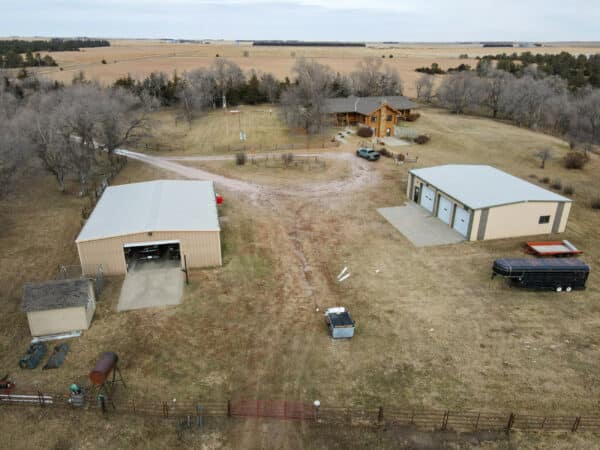 Aerial view of a rural ranch for sale featuring a house, two large metal outbuildings, a shed, trailer, and scattered vehicles, all surrounded by open fields and trees under a cloudy sky.
