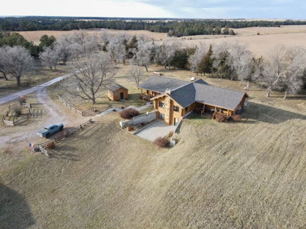 Aerial view of a large wooden cabin with a sloped yard, surrounded by trees and open fields. Ideal as a ranch for sale or hunting property, with a driveway leading to the house, a truck parked nearby, and a small shed to the left.
