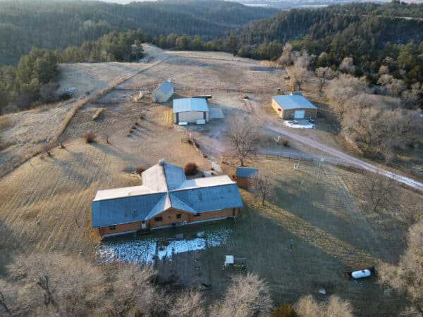 Aerial view of a rural hunting property with a large house, several outbuildings, patchy snow, open fields, trees, and a dirt driveway winding through the landscape. Ideal land for sale for those seeking privacy and outdoor adventure.
