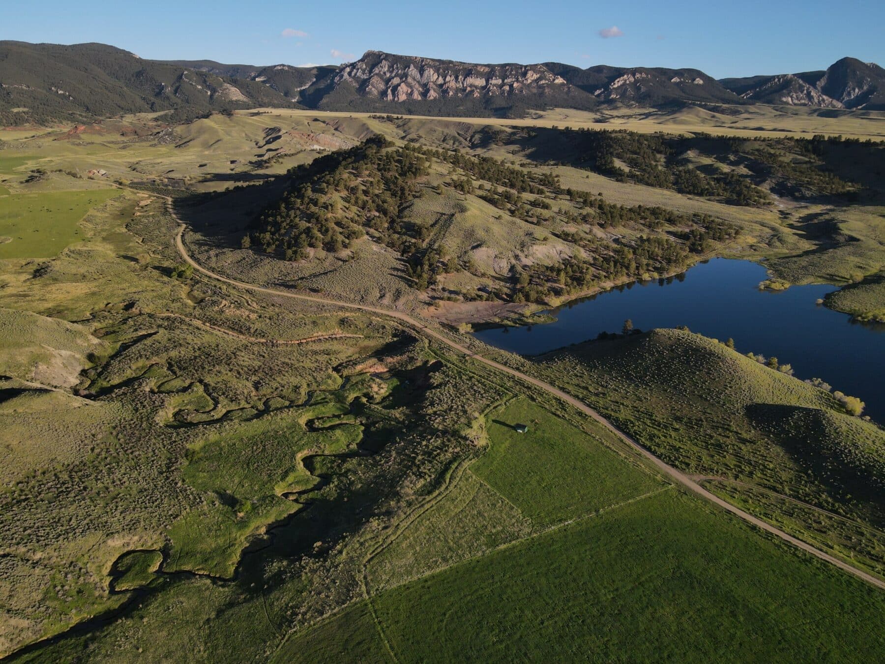 Aerial view of a green valley with rolling hills, a winding dirt road, and a blue lake, set against rocky mountains under a clear blue sky—perfect recreational land or ranch for sale.