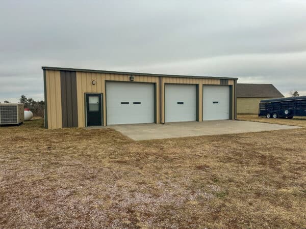 A metal building with two large garage doors and a single entry door sits on a concrete pad, surrounded by grassy land for sale under an overcast sky. A trailer and another building are visible in the background.
