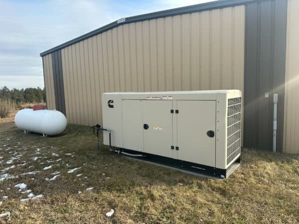 A large backup generator is installed next to a beige metal building on a grassy area with patches of snow—an ideal feature for recreational land or a cattle ranch. A white propane tank is visible nearby.