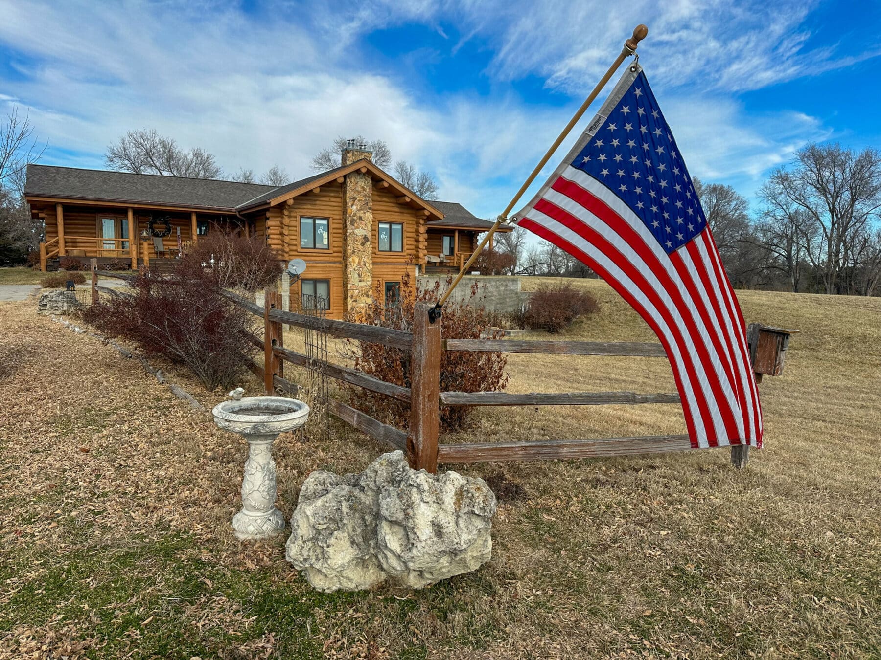 A large American flag hangs on a wooden fence in front of a log cabin house, surrounded by dry grass and leafless trees under a partly cloudy blue sky—a perfect scene for a hunting property or potential ranch for sale. A stone birdbath stands nearby.