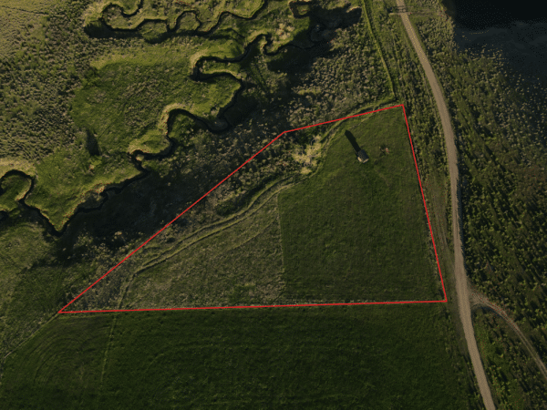 Aerial view of a triangular grassy field outlined in red, bordered by a road and winding water channels—ideal as recreational land or hunting property; a small structure stands near the center of the area.