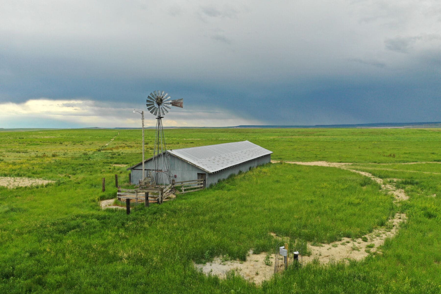 A metal windmill and a long, gray barn sit alone in a vast green field under a cloudy sky, with patches of sand and distant horizon—perfect scenery for a cattle ranch or land for sale.