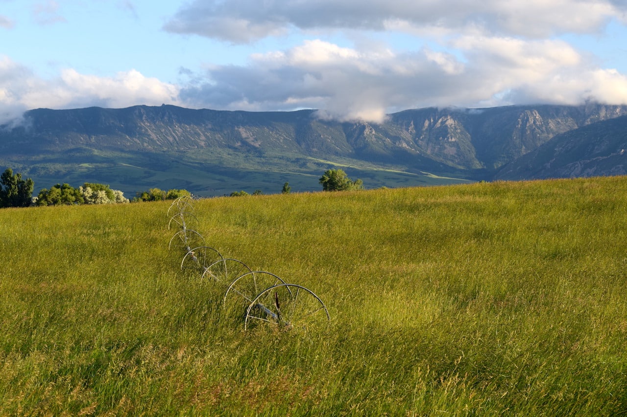 A grassy field with old irrigation wheels stretches toward distant tree-covered mountains under a partly cloudy sky, ideal as recreational land or a scenic cattle ranch.