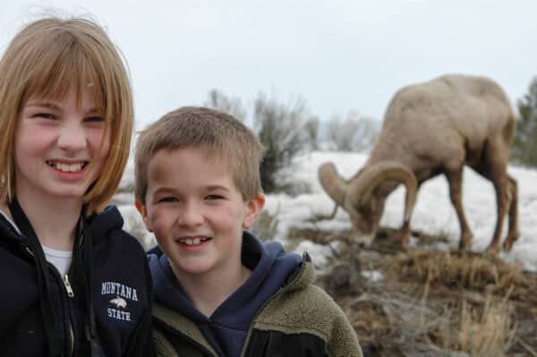Two smiling children stand outdoors in winter jackets on recreational land, with a large bighorn sheep grazing in the snowy background. The scene highlights the natural beauty of a ranch for sale in a cold, scenic setting.