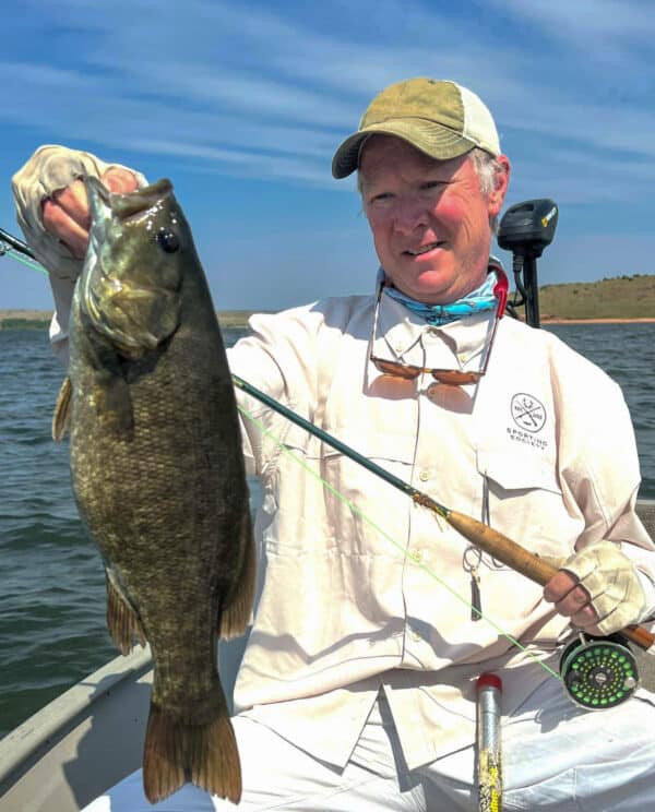 A person in a boat holds up a large fish with one hand while holding a fishing rod in the other. The person is smiling and wearing a light shirt, gloves, and cap, enjoying the water and shoreline—perfect for recreational land adventures.