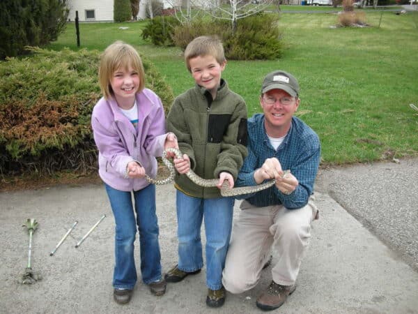 Two children and an adult crouch outside on pavement, smiling and holding a long snake together. Green bushes and grass are in the background, suggesting a hunting property or cattle ranch setting. The children wear jackets and jeans; the adult wears a cap and glasses.