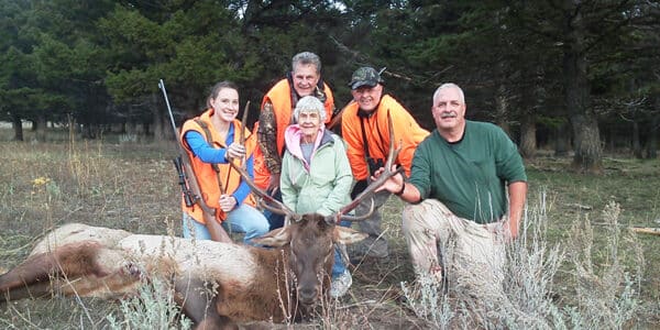 Five people pose outdoors in a grassy, wooded area on a hunting property behind a large elk lying on the ground. Three wear orange hunting vests and hold rifles. All are smiling at the camera, with trees filling the background.