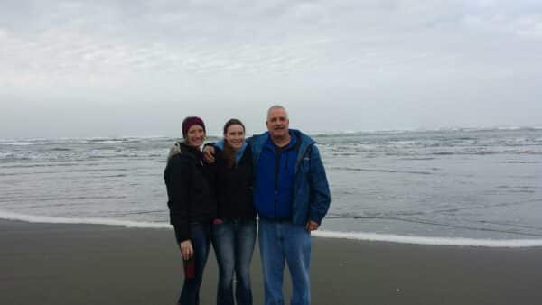 Three people stand close together and smile on a cloudy beach with gentle waves behind them. They wear jackets and jeans, suggesting cool weather. The ocean and cloudy sky stretch out, evoking the open feel of recreational land for sale.