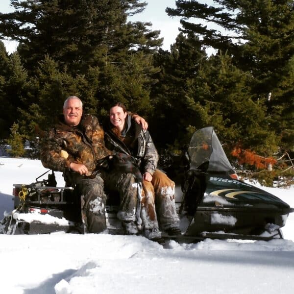 Two people dressed in winter camouflage sit together on a snowmobile in a snowy forest, smiling at the camera with evergreen trees in the background—perfect scenery for recreational land or hunting property.