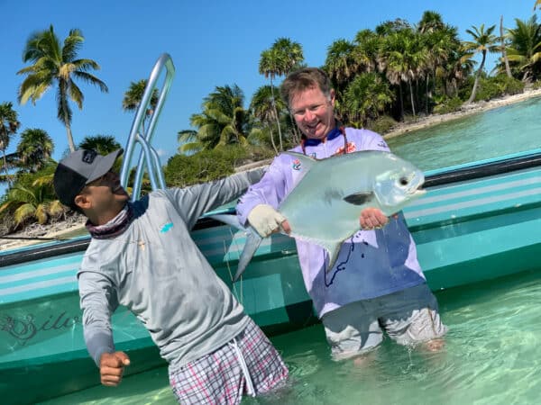 Two men stand in shallow tropical water next to a boat; one smiles while holding a large silver fish as the other laughs. Palm trees and a sandy beach are in the background—perfect for those seeking recreational land or a ranch for sale.