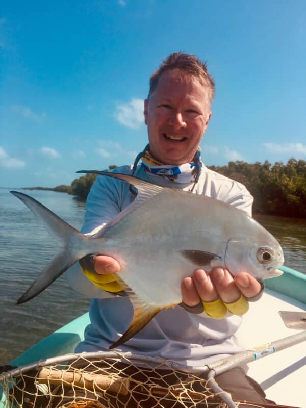 A man sitting in a small boat holds a silver fish with both hands, smiling at the camera. Behind him, calm water and greenery stretch under a blue sky—a perfect day for fishing or exploring nearby hunting property or land for sale.