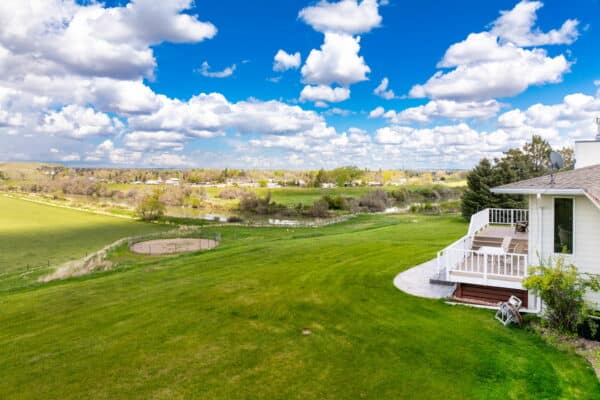 A large, green lawn stretches toward a distant river and scattered trees under a blue sky with fluffy clouds; a white house with a deck is visible on the right side, perfect for recreational land or as part of a scenic ranch for sale.