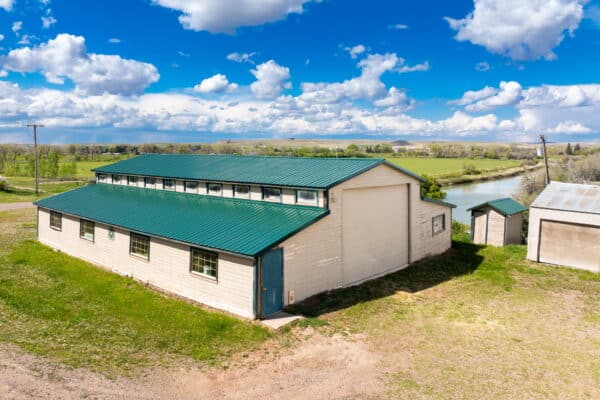 A large white building with a green metal roof stands near a small river on recreational land, surrounded by grassy fields and scattered trees under a blue sky with fluffy clouds.