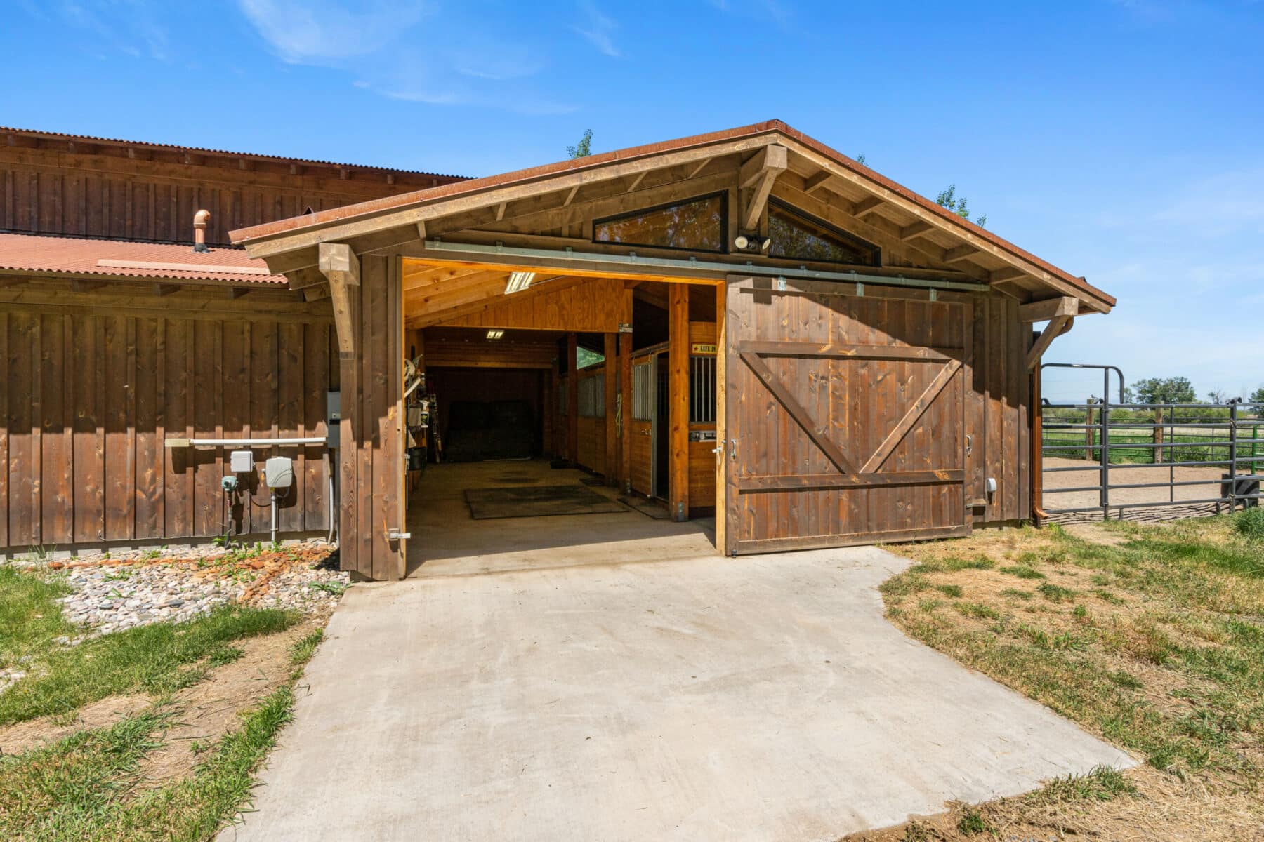 A wooden barn with an open sliding door reveals a clean interior. Set on a cattle ranch, it’s surrounded by grass, a concrete driveway, and a fenced area under a bright blue sky—perfect land for sale.