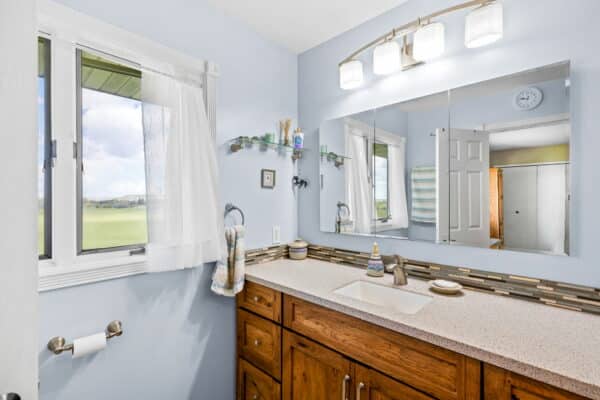A bright bathroom with a large mirror, wooden vanity, single sink, and modern light fixture. Natural light streams in through a window with sheer curtains. Towels hang by the sink, and the countertop holds toiletries—a peaceful retreat for your cattle ranch home.