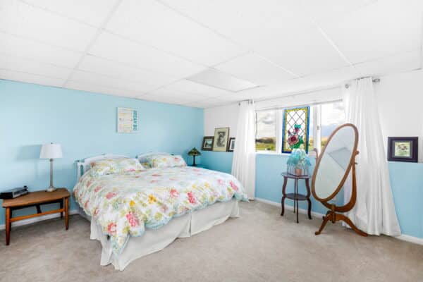 A cozy bedroom with a floral-patterned bed, blue and white walls, a stained-glass window, a standing mirror, and simple decor makes this ranch for sale inviting and comfortable. Framed pictures and a nightstand with a lamp complete the space.