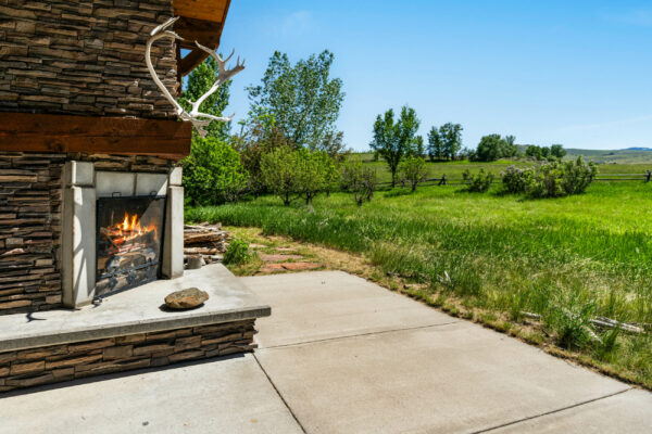 A stone outdoor fireplace with a lit fire and mounted antlers stands on a patio overlooking recreational land, grassy yard, trees, and a wooden fence under a clear blue sky.