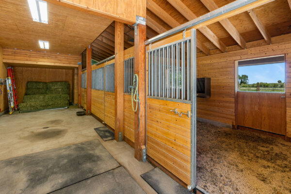 A clean, well-lit horse stable with wooden stalls and metal bars, hay bales stacked at the back, and a window overlooking a grassy field—perfect for those seeking ranch for sale or recreational land. Rubber mats cover parts of the floor.