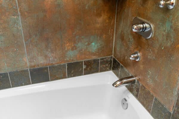 Close-up of a white bathtub with a metal faucet and bath controls, set against rustic earth-toned tiles in shades of brown, green, and copper—an inviting retreat perfect for relaxing after a day on your dream ranch for sale.