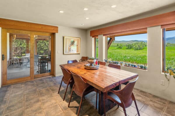 A dining room with a rustic wooden table and six chairs, large windows overlooking a green field and mountains, potted plants on the windowsill, and sliding glass doors leading to a patio—perfect for enjoying life on a cattle ranch for sale.