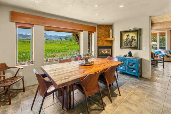 A bright dining room with a wooden table, eight chairs, tile flooring, and large windows overlooking a green field and mountains—perfect for a hunting property. Features a corner fireplace, rustic decor, and an adjacent living area.