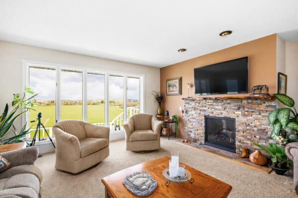 Bright living room with large windows overlooking recreational land, featuring tan armchairs, a stone fireplace with a TV above, houseplants, and a wooden coffee table with decor items.
