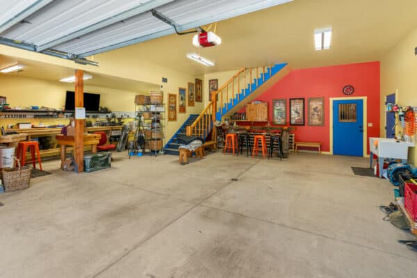 Spacious, organized garage with workbenches, tools, stools, and art on yellow walls. Blue stairs lead to a loft. Open garage door lets in light; various items are neatly arranged—perfect for a ranch for sale or recreational land setup.