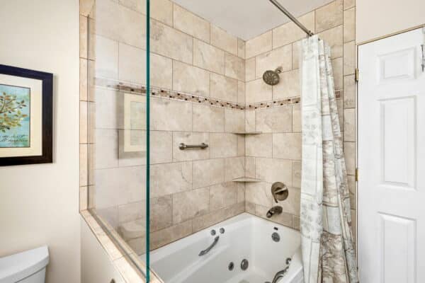 A bathroom with a beige tiled shower and bathtub combo, featuring a showerhead, built-in shelves, grab bar, and a white shower curtain with map designs—perfect for relaxing after exploring your new cattle ranch. A framed picture hangs on the wall next to the tub.