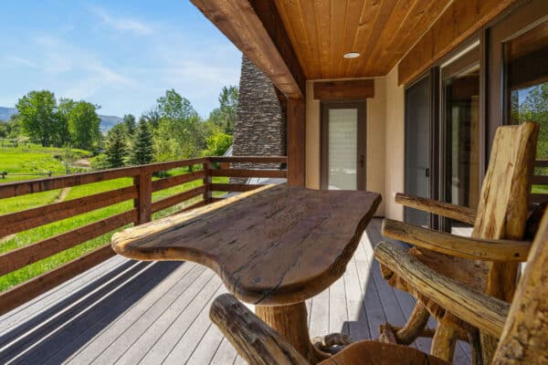 A rustic wooden table and chairs sit on a covered deck with railings, overlooking lush recreational land with trees and hills under a clear blue sky—perfect for enjoying a hunting property or exploring this unique ranch for sale.
