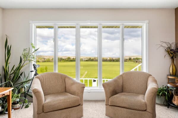 Two beige armchairs sit in front of large windows overlooking a green field, hinting at expansive land for sale. Indoor plants and a small side table decorate the cozy, carpeted room, with natural light streaming in from the windows.