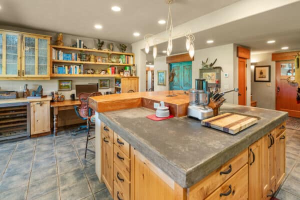 A spacious kitchen with a central island featuring a concrete countertop, wooden cabinets, a food processor, and a desk area with bookshelves and a computer—perfect for life on a cattle ranch. Warm lighting and tile flooring add to the inviting atmosphere.