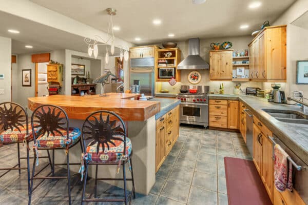 Southwestern-style kitchen with wood cabinets, a large island with three decorative chairs, stainless steel appliances, colorful tile accents, and pendant lighting on blue-gray tiled flooring—perfect for a ranch for sale or spacious cattle ranch home.