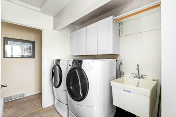A laundry room with a front-loading washer and dryer, white wall cabinets above, utility sink, wooden rod for hanging clothes, and white walls. The doorway opens to a carpeted area—perfect for a cattle ranch or hunting property.