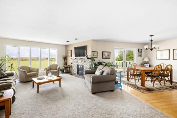 Bright living and dining area with large windows, beige carpet and sofa, armchairs, coffee table, stone fireplace with TV, indoor plants, and a wooden dining table on a rug. Enjoy views of the green lawn on this beautiful cattle ranch land for sale.