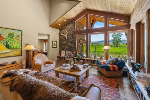 A cozy living room with large windows overlooking scenic recreational land, featuring leather armchairs, a fur throw, wooden table, eclectic decor, bookshelves, and natural light highlighting rustic charm.