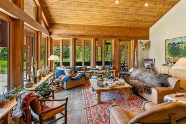 Cozy living room with wooden walls and ceiling, large windows overlooking greenery, sofas draped with colorful blankets, a coffee table, plants, books, and eclectic decor—perfect for relaxing after a day exploring your hunting property or cattle ranch.