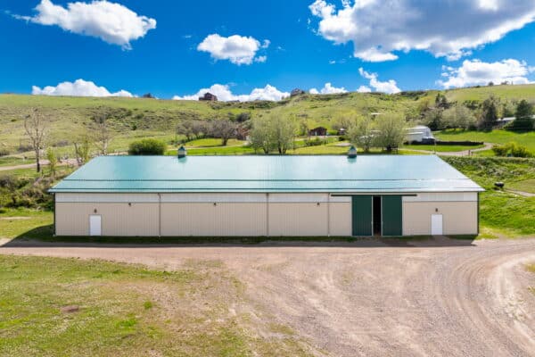 A large white barn with a green roof stands on a dirt lot, perfect for recreational land or a cattle ranch, surrounded by grassy hills and scattered trees under a bright blue sky with scattered clouds.