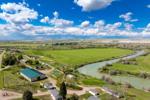 Aerial view of a green rural landscape with fields, scattered houses, a curving river, trees, and a bright blue sky—ideal as recreational land or a picturesque cattle ranch for sale.