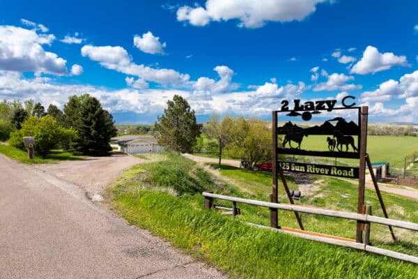 A rural road leads to a white house and garage, surrounded by trees and green fields. A sign reading 2 Lazy C, 125 Sun River Road, with horse silhouettes marks this scenic ranch for sale under a bright blue sky with clouds.