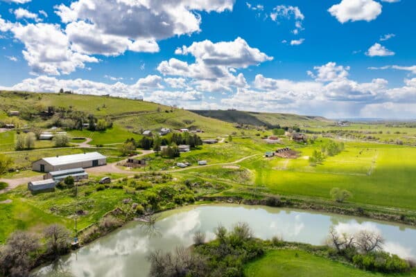 A scenic rural landscape with green fields, scattered buildings, hills, and a reflective pond under a partly cloudy blue sky—ideal recreational land or cattle ranch available.
