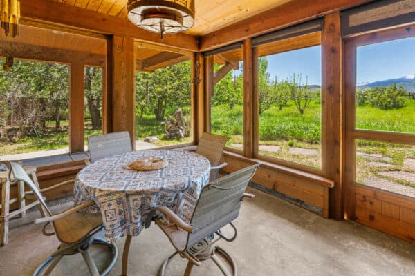 A cozy screened-in porch with a round table covered by a patterned tablecloth, four chairs, wooden beams, and large windows overlooking green yard and distant mountains—perfect for relaxing after exploring your ranch for sale or nearby hunting property.