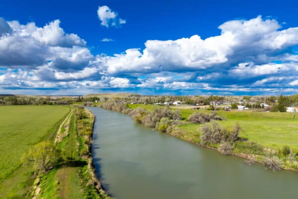 A wide river flows through green fields and trees under a partly cloudy blue sky, with land for sale near a small town and hills visible in the background.