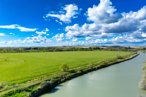 A wide river curves alongside a vast green field under a bright blue sky filled with white, fluffy clouds. This open and peaceful landscape, with patches of trees and distant hills, would make an ideal cattle ranch or recreational land.