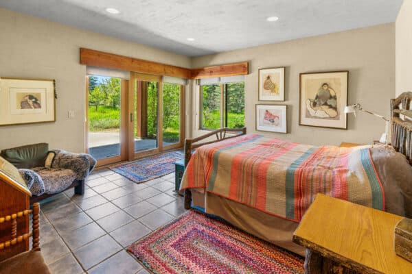 A cozy bedroom with large glass doors opening to greenery on recreational land, a bed with a colorful patchwork quilt, framed art on beige walls, a wooden desk, armchair, and woven rugs on a tiled floor.
