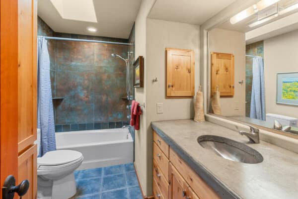 A bathroom with a blue-tiled floor, concrete countertop and sink, wooden cabinets, toilet, and bathtub with shower and blue curtain. Two decorative vases and a towel sit on the counter—a peaceful retreat perfect for ranch for sale or recreational land living.