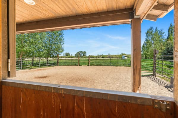 View from inside a wooden stable looking out onto a fenced, sandy corral with green grass, trees, and a clear blue sky—ideal for those seeking a cattle ranch or land for sale.